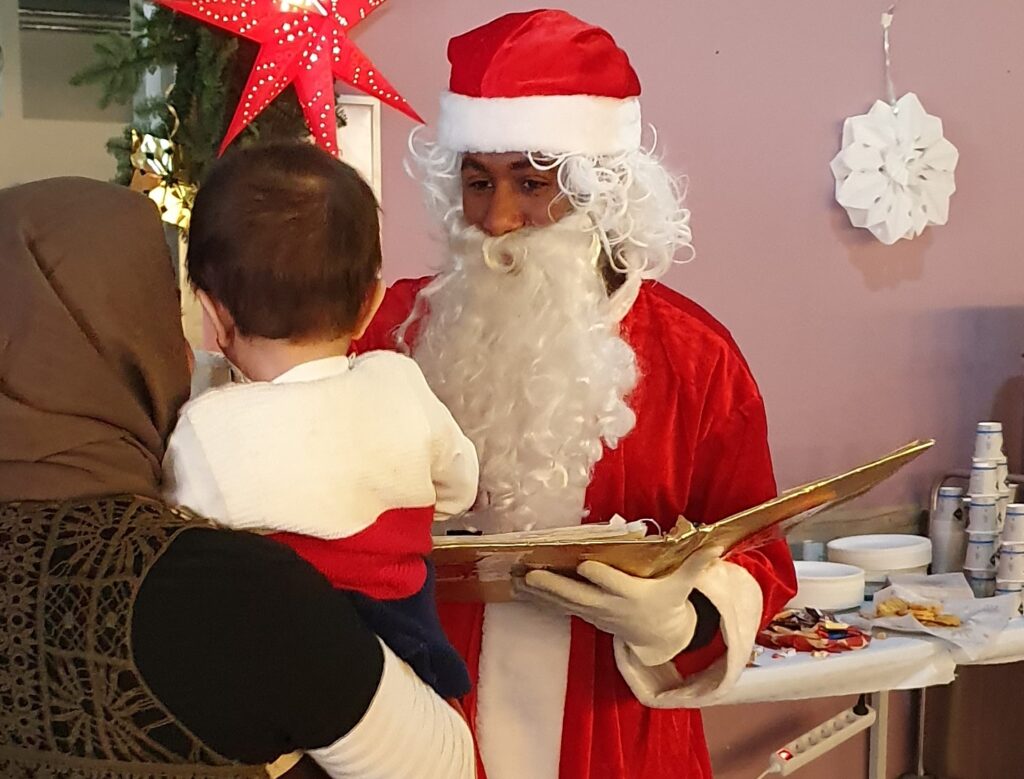 Ein als Weihnachtsmein verkleideter junger Mann übergibt ein Geschenk an ein Kleindkind, das auf dem Arm der Mutter gehalten wird